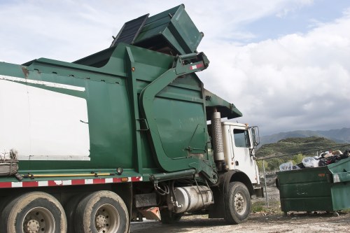 A large industrial waste collection truck operated by Hampstead Heath House Clearance is positioned on a gravel driveway, actively tipping a green plastic skip filled with mixed rubbish. The truck features a white cab with a metallic finish and a sturdy, reinforced chassis with six large black tires, three on each side. The skip, which is depicted in a raised position at the rear of the vehicle, has a textured surface and visible wear, indicating frequent use. To the right of the truck, a green wheelie bin brimming with assorted waste materials, including black and white rubbish bags and other debris, is situated on the uneven ground. The background showcases cloudy skies with patches of blue, rugged hills, and distant landscape typical of the Hampstead Heath area in North London. The scene emphasizes the practical aspect of rubbish removal services in the region, providing clear visual context of waste collection and disposal operations carried out by Hampstead Heath House Clearance.