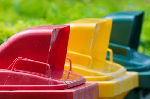 A row of four large outdoor waste bins positioned on a paved surface in a residential area, with a background of green grass and blurred foliage. The bins are made of durable plastic with textured surfaces and have hinged lids, each in a different color: red, yellow, blue, and green. The red bin is closest to the camera, followed by the yellow, blue, and green bins, arranged in a straight line. The lids are closed, and the bins appear to be clean and well-maintained, suitable for rubbish collection or waste disposal services such as those provided by Hampstead Heath House Clearance in North London. The setting suggests an organized outdoor space, possibly near a driveway or pavement, ready for rubbish removal or house clearance tasks typical in Hampstead Heath or surrounding areas.